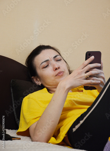 A young, beautiful woman is lying on the sofa with a phone in her hands. She is looking at the smartphone screen and frowning with dissatisfaction. negative emotions are visible on her beautiful face