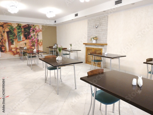 The dining area of a cafe or canteen, featuring several rectangular tables and chairs. Small vases of flowers, napkins, and cutlery are placed on the tables.