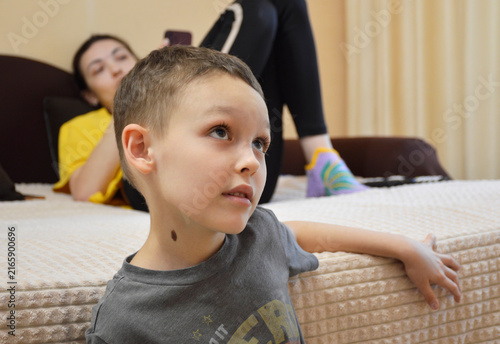 young boy in a gray T-shirt is sitting on the edge of a bed or mattress. He is looking off to the side with a pensive expression. In the background, a woman is lying on the bed, holding a smartphone.