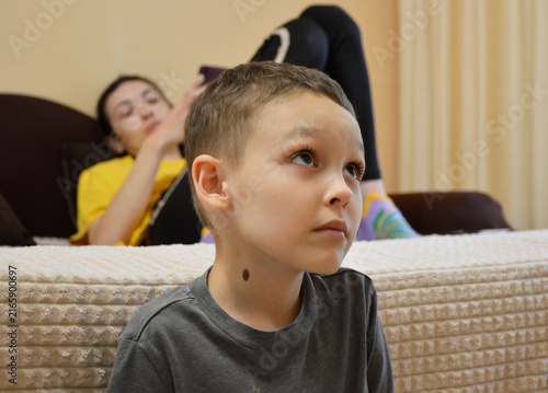 young boy in a gray T-shirt is sitting on the edge of a bed or mattress. He is looking off to the side with a pensive expression. In the background, a woman is lying on the bed, holding a smartphone.