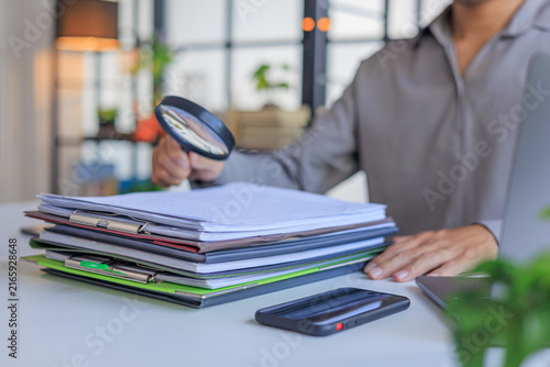 Bild auf Leinwand Professional closely inspecting a stack of organized documents and file folders on a desk with a magnifying glass, symbolizing a detailed financial audit, document review, or fraud investigation