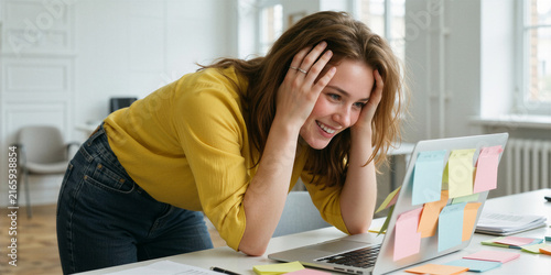 Overwhelmed young woman with optimistic smile sits at white desk, clutching her face, surrounded by colorful sticky notes in a bright modern office.