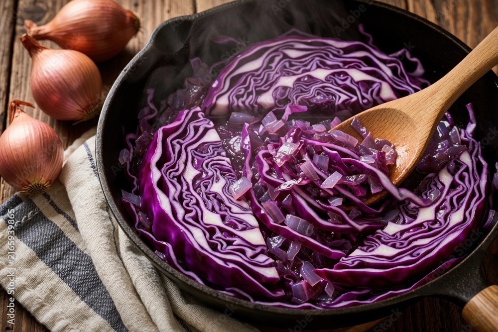 Fototapeta premium Red cabbage cooks in cast iron pan with onions and wooden spoon. Steam rises from the vegetable dish prepared for dinner on rustic wooden table.