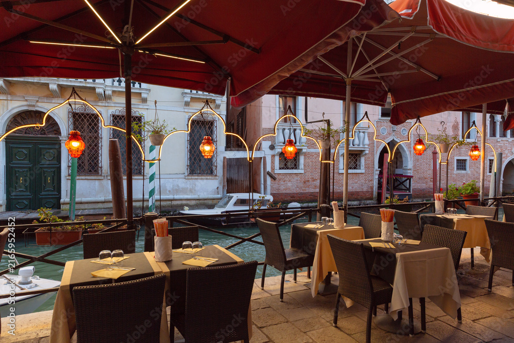 Fototapeta premium Outdoor restaurant terrace beside a narrow Venetian canal with tables, boats and traditional architecture in Venice, Italy.