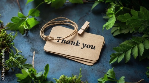 Expressing gratitude with a paper tag featuring thank you text surrounded by lush green foliage and fresh leaves on a rustic dark textured surface creating an elegant organic composition