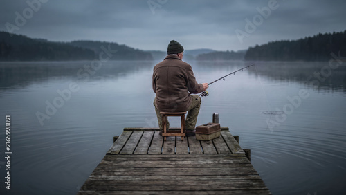 A solitary man in a brown jacket sits on a wooden dock fishing in a misty, calm lake. A tackle box and thermos rest nearby in the tranquil, overcast morning.