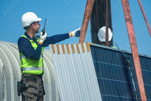 Canvastavla Two renewable energy specialists in hard hats and hi vis workwear precisely direct the crane over the module stack by radio high on an industrial warehouse roof in a Thai city