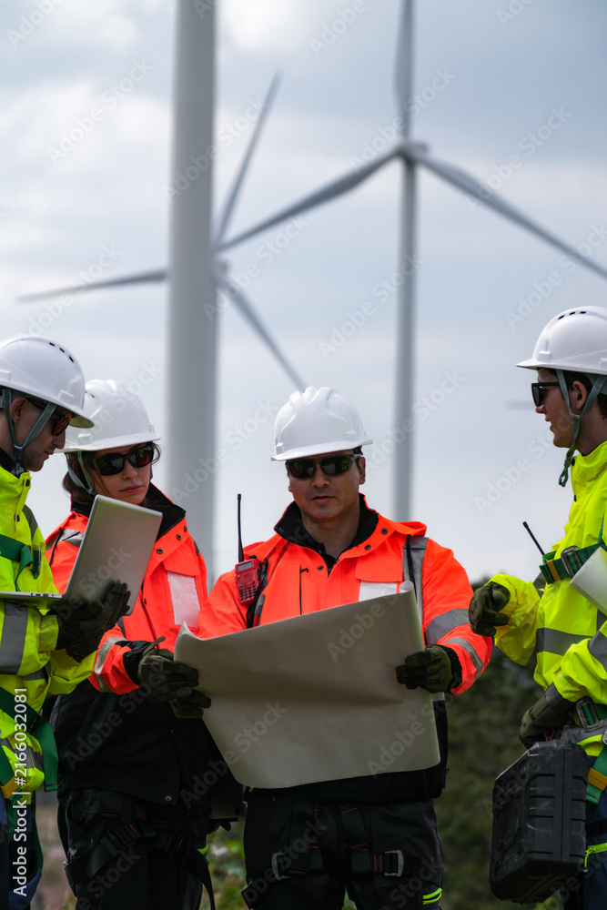 Fototapeta premium Wind project engineers with laptop, radio and drawings coordinate with the control room by radio gathered on open ground beneath the turbines in silver overcast light at an operating.