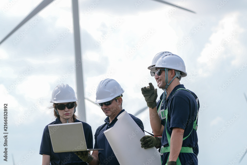 Fototapeta premium A four person rigging crew in navy suits and white helmets debate the lift plan while carrying the load under turning rotors on the wind farm in bright hazy daylight at an operating.