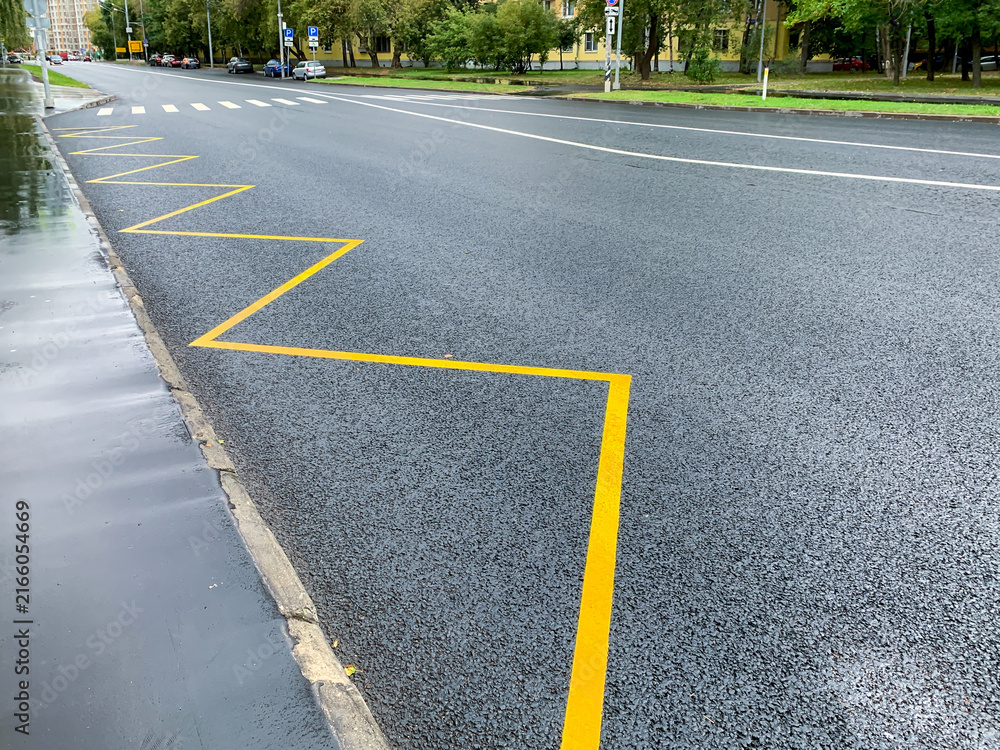 Obraz premium A freshly renewed yellow zigzag marks the bus stop zone along the kerb of a wet city street. New asphalt and white lane lines shine after the rain. Urban road infrastructure