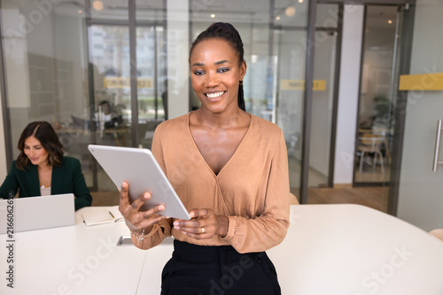 Fotografía Portrait of successful African businesswoman holds tablet, reviewing project presentation, female colleague works on laptop in background