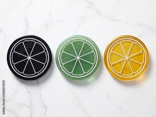 Three colorful round glass coasters with citrus fruit slice patterns on a white marble surface, top view