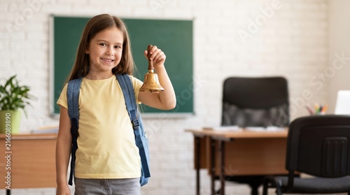 Happy little girl holding a school bell in a classroom, ready for the first day of school.