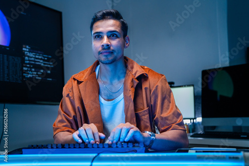 Fotografía A dark-haired network defence analyst concentrates at the keyboard in the dark room in a dimly lit network monitoring room, surrounded by glowing monitors and code