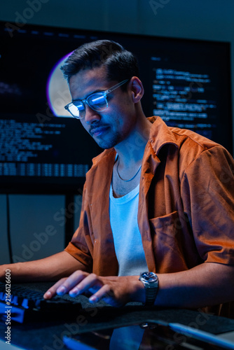 Photography A focused dark-haired cybersecurity analyst works front-facing at the monitor in a blue-lit cyber operations centre, monitoring live data streams for cyber threats and intrusions