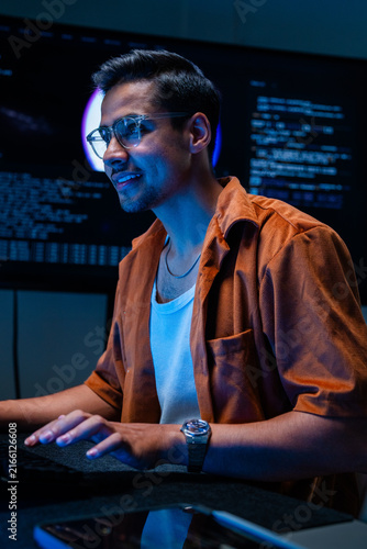 Fotografie A dark-haired network defence analyst concentrates at the keyboard in the dark room in a shadowy data monitoring room lit by screen glow, defending the corporate network