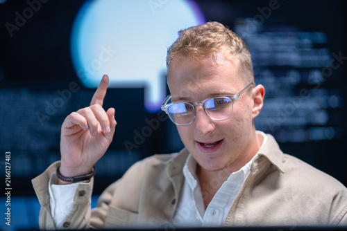 A fair-haired security engineer leans toward the display reading security alerts in a shadowy data monitoring room lit by screen glow, surrounded by glowing monitors and code.