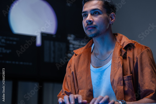 Fotografía A dark-haired network defence analyst pauses in thought at his desk at a multi-monitor workstation in a darkened control room, surrounded by glowing monitors and code