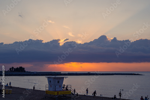 Bild auf Leinwand Beach at Sunset with Lifeguard Tower, Swimmers, Dramatic Clouds and Crepuscular