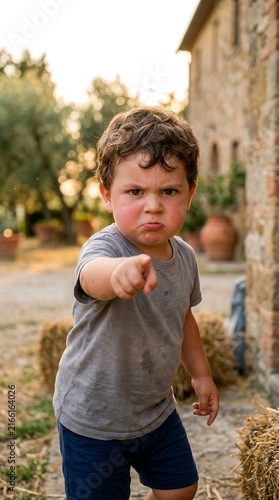 Young boy with a grumpy expression pointing his finger directly at the camera outdoors