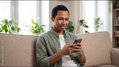 Smiling happy young Indonesian man holding cell mobile phone using smartphone sitting at home on couch, scrolling social media, checking financial apps, buying online, texting messages. 