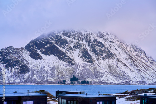 Majestic winter mountain peak glowing in spectacular golden hour light in Lofoten, Norway. Beautiful scenic view of rugged snow-covered Arctic mountains illuminated by warm sunrise or sunset under a d