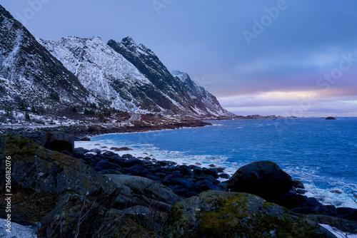 Majestic winter landscape of Lofoten islands in Norway with bright sunlight reflecting on the calm blue sea. Beautiful scenic view of grand snow-covered mountains under a bright dramatic sky in Arctic