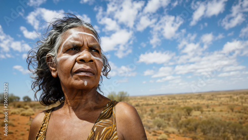Portrait of a wise senior Aboriginal woman with traditional white paint on her face, standing in the vast Australian outback under a bright blue sky with white clouds.
