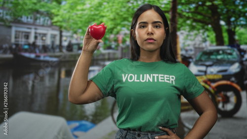 Fotografía Young hispanic woman volunteer in green shirt holds small red heart aloft with raised hand on a street; compassion hope solidarity