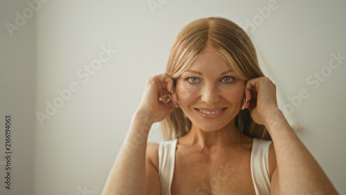 Canvastavla Woman touching her face with hands in a bright studio, smiling while adjusting a white tanktop strap at the neckline and touching her cheek; serenity