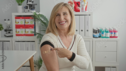 Cuadro en lienzo Woman pointing finger to band aid on upper arm in medical building, smiling after vaccination and showing post injection care; health confidence