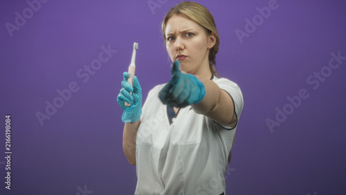 Bild auf Leinwand Woman dentist wearing white uniform and blue gloves points finger to camera while holding toothbrush in studio; stern warning