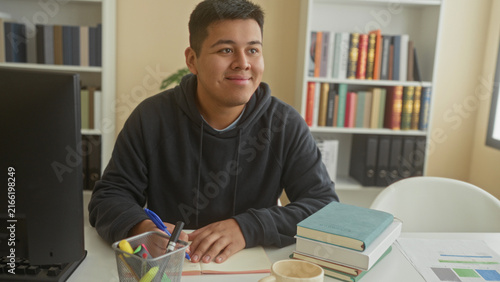 Canvastavla Man writing with pen at desk in a building library, smiling while taking notes on paper and referencing stacked books; concentration learning