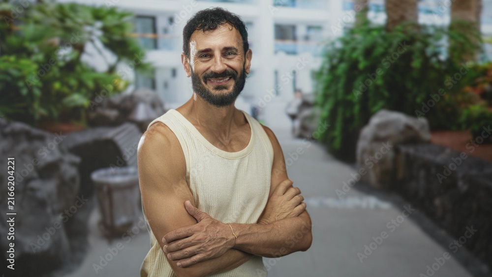 Fototapeta premium Man standing with arms crossed showing muscular forearms and smile, wearing sleeveless top in a hotel building courtyard; confidence.