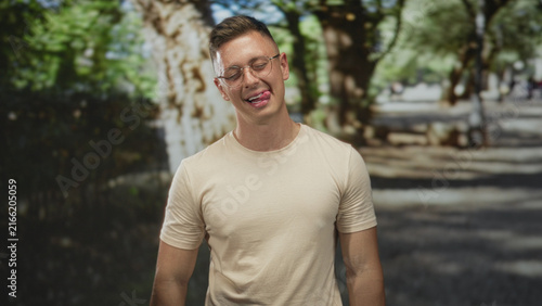 Man sticking tongue out and smiling, glasses visible, beige tee on sunlit tree lined park walkway with trees and trunk nearby; playful.