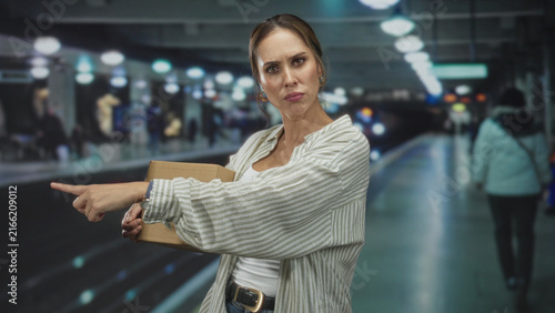 фотографія Woman holding cardboard box points finger forward in building transit platform with assertive gaze and outstretched arm; delivery determination