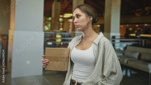 Fotografie Woman holding a cardboard box with her right hand at chest, pouting and glancing upward, wearing white tank and open striped shirt, standing in a building lobby; frustration waiting delay