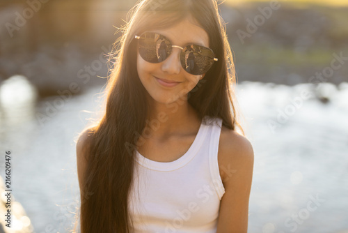 At a serene riverside during golden hour, a young girl with long hair flashes a joyful smile. She wears fashionable sunglasses that mirror the peaceful scenery around her