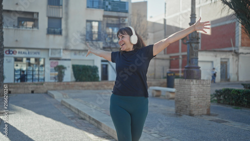 Konstfotografi Woman with white headphones and arms outstretched smiling on street in front of buildings, clearly dancing and enjoying music; joyful freedom