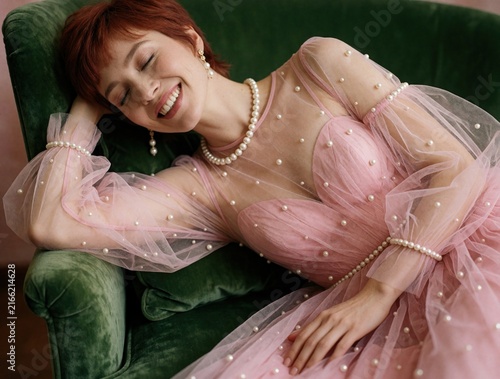 Smiling young woman with short red hair wears pink tulle dress with pearl details. Beautiful female model reclines on green velvet armchair. Elegant lady enjoys luxury lifestyle  feeling joyful 