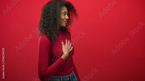Fotografie Teenage woman places hand to chest in red sweater against studio wall, side profile and subtle smile; sincerity introspection