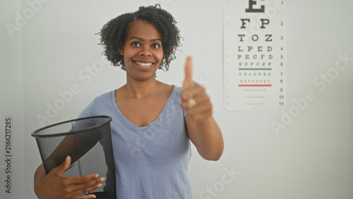 Foto Woman optician smiling throws glasses into mesh trash bin with hand beside eye chart in clinic building; empowerment relief