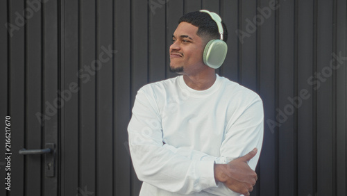 Man wearing green headphones smiling with arms crossed while looking aside  standing by a black metal building door  urban calm contentment.