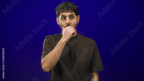 Fotografie Man covering mouth with hand in studio blue backdrop, wearing brown tshirt and earring; surprise silence