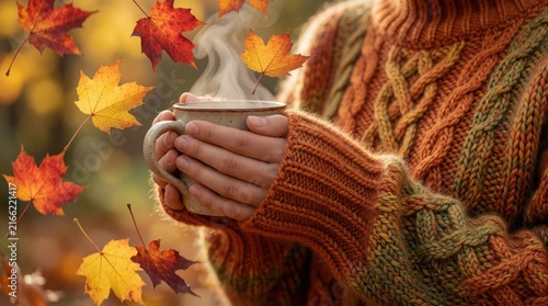 Person in warm orange knitted sweater holds steaming ceramic mug outdoors. Golden autumn leaves fall around hands with hot tea. Cozy seasonal lifestyle concept with rustic fall vibes, peaceful nature