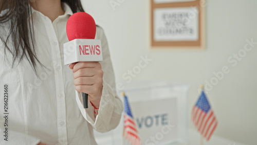 Photography Woman holds news microphone with visible hand beside a vote sign at a polling station inside a building; patriotic civic duty