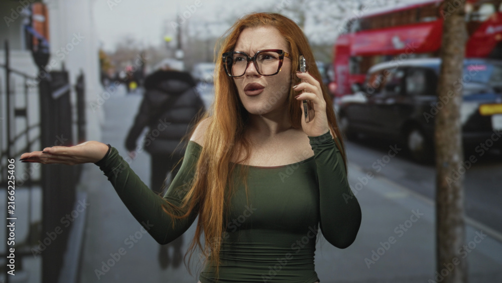 Fototapeta premium Redheaded woman on a street holding a smartphone to her ear with palm raised in an exasperated shrug, wearing glasses and green off shoulder top; frustration confrontation.