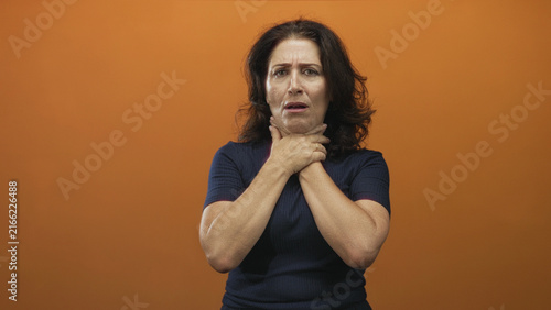 Quadro su tela Middle aged woman clutching neck with both hands in studio orange backdrop, choking gesture and tense expression; panic urgent help