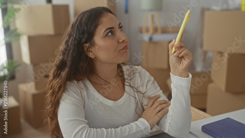 Принти на полотні Woman holding a yellow highlighter to her lips in a building surrounded by cardboard boxes and a notebook while thinking about move details; quiet contemplation