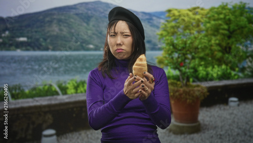 Картината върху платно Woman in purple turtleneck holds croissant with a grimace, pushes hand away and shrugs slightly on lakeside terrace; disgust rejection